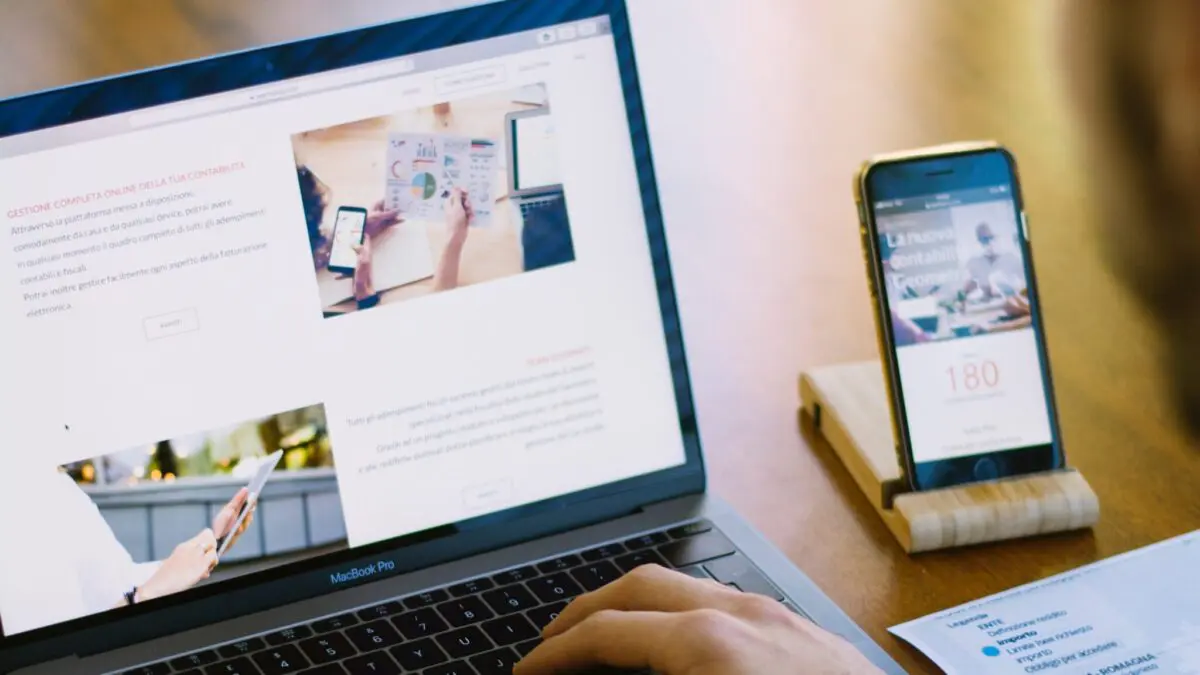 Person developing a website on a laptop, with the same site open on a mobile device on the table beside them.